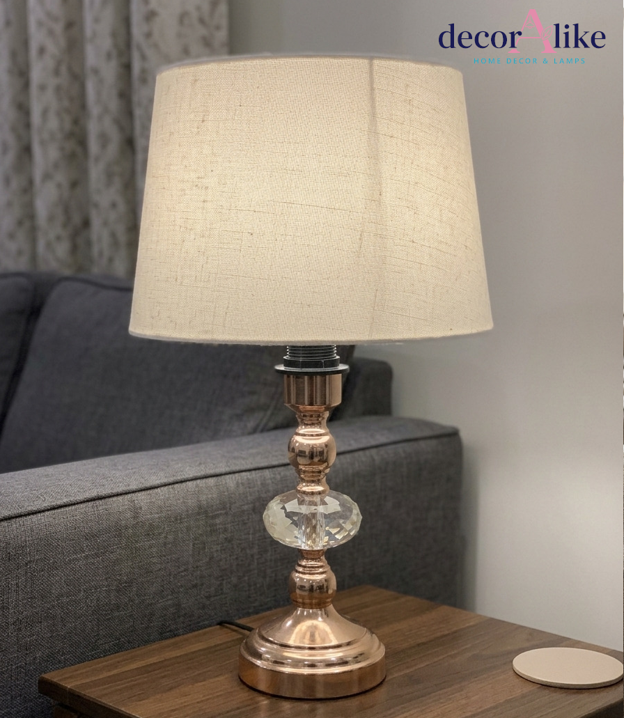 Rose gold table lamp with faceted crystal accent and beige linen shade on a wooden bedside table