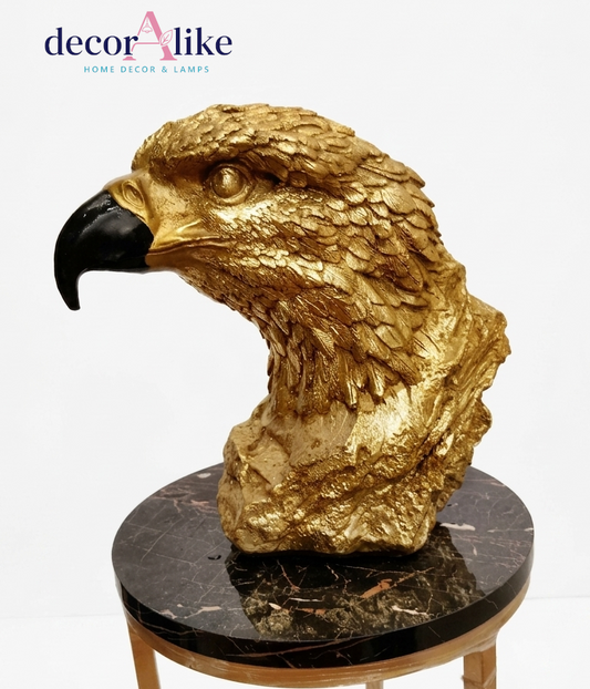 A golden eagle head sculpture with a black beak, displayed on a round black marble-topped table against a white background. The 'decorAlike' brand logo is visible in the top left corner.