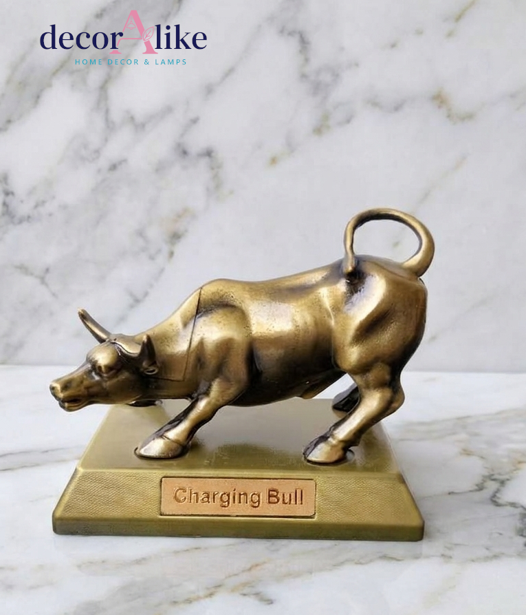 A bronze-finished charging bull figurine standing on a rectangular base engraved with 'Charging Bull'. The product is photographed on a white marble surface with a matching marble background, and the 'decor A like' brand logo is visible in the top left corner.