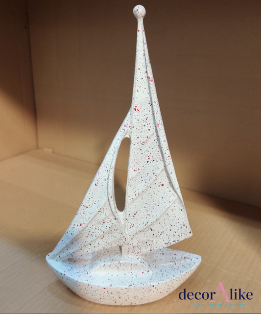 A white speckled decorative sailboat candle holder with a central cutout in the sail, displayed on a cardboard shelf. The product features red and black speckles on a white base.