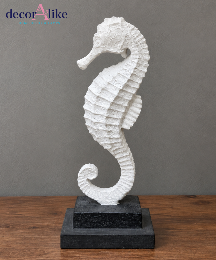 A white seahorse figurine sculpture standing on a two-tier black base, set against a textured gray wall and a wooden tabletop. The 'decorAlike' brand logo is visible in the top left corner.