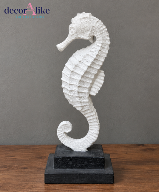 A white seahorse figurine sculpture standing on a two-tier black base, set against a textured gray wall and a wooden tabletop. The 'decorAlike' brand logo is visible in the top left corner.
