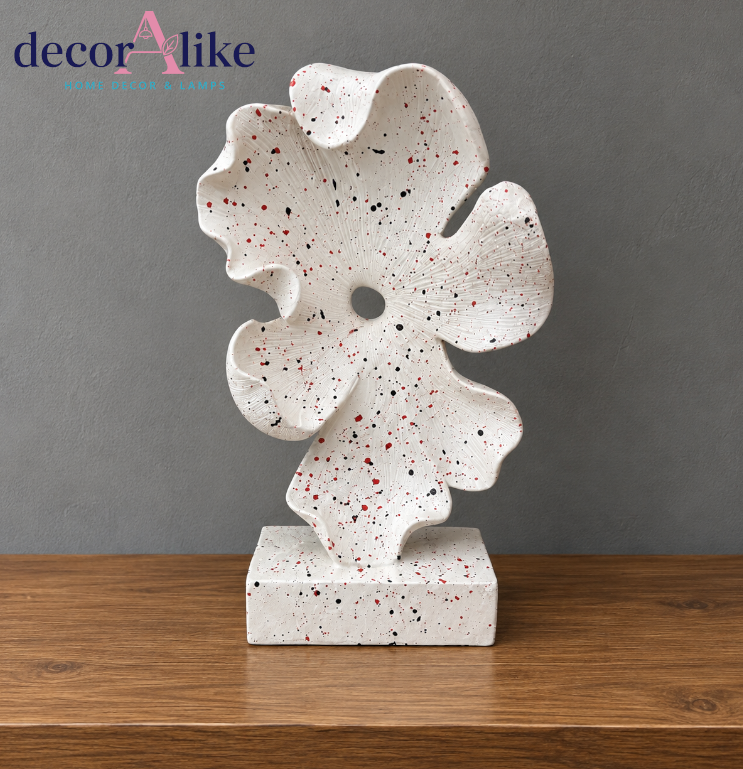 An abstract sculptural tabletop decor piece with a white stone-like finish and red, black, and gray speckles, displayed on a wooden surface against a gray wall. The brand logo 'decorAlike' is visible in the top left corner.