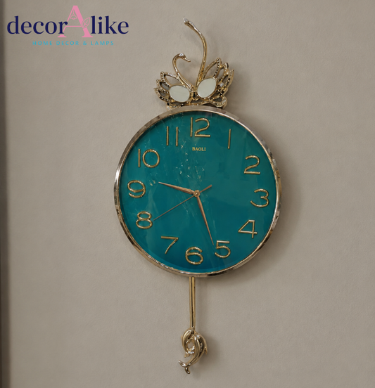 A decorative wall clock with a round turquoise blue face, gold-tone numerals and hands, a gold-tone top scroll ornament, and a dangling gold-tone bottom ornament, shown against a plain beige wall. The brand name 'MADE' is visible on the clock face.