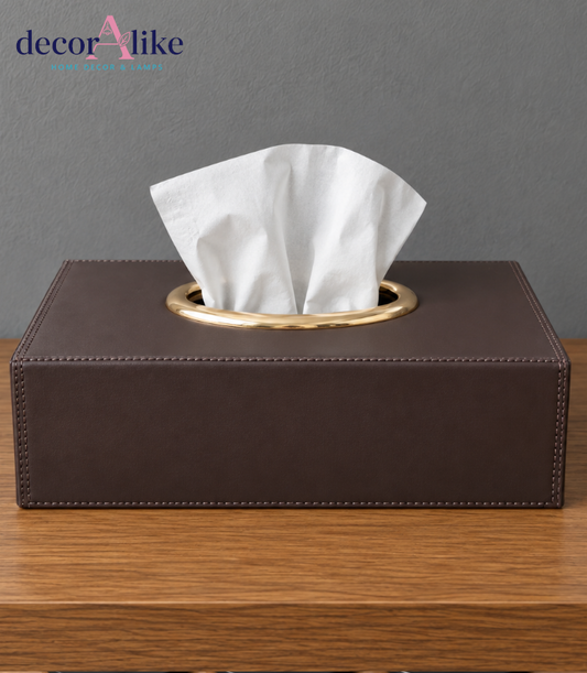 A dark brown faux-leather tissue box cover with a gold metal rim and a white tissue paper pulled out, sitting on a wooden table against a gray wall. A 'decorAlike' watermark is visible in the top left corner.