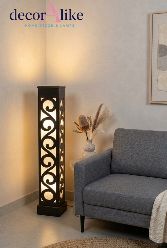 A tall black metal floor lamp with an intricate scrollwork cutout pattern, glowing with warm light in a styled living room corner next to a gray armchair.