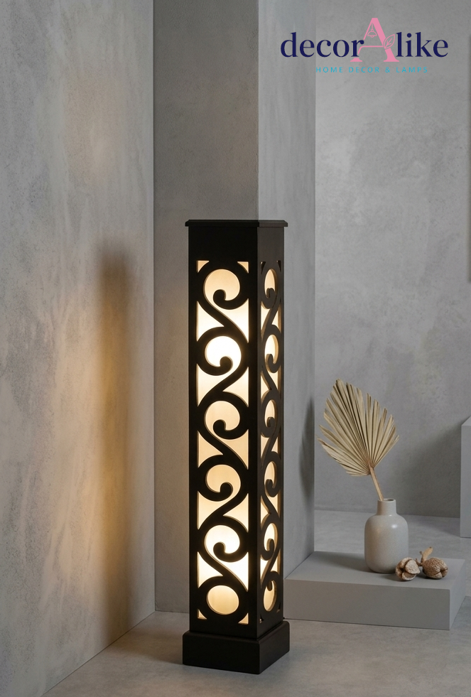 A tall rectangular outdoor-style floor lamp with a black metal frame and illuminated scrollwork cutouts, placed in a modern interior setting with textured gray walls and a small vase with dried leaves nearby. The decorAlike brand logo appears in the top right.
