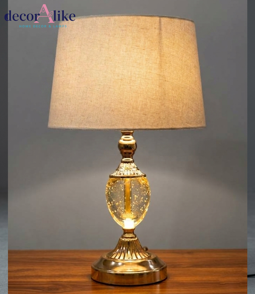 Gold table lamp with clear seeded-glass body and beige fabric shade on a wooden table