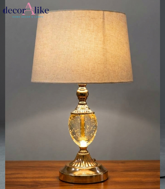 Gold table lamp with clear seeded-glass body and beige fabric shade on a wooden table