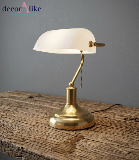 A brass bankers lamp with a white curved glass shade, resting on a rustic wooden table against a gray wall. The brand logo 'decorAlike' is visible in the top left corner.