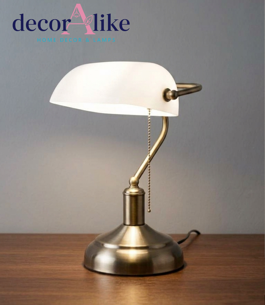 A vintage-style banker's desk lamp sits illuminated on a smooth wooden tabletop against a plain gray wall. The lamp features a classic, rounded white glass shade, a curved antique brass or bronze neck, and a matching rounded metallic base. A beaded metal pull-chain hangs straight down from underneath the white glass shade, which glows softly from the light bulb inside.
