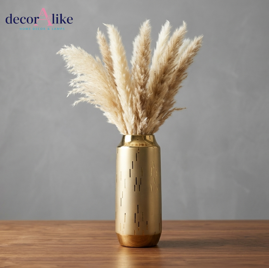 A gold cylindrical vase with a brushed metallic finish and small vertical cutout details, holding dried pampas grass, set on a wooden surface against a textured gray wall. The decorAlike brand logo is visible in the top left corner.