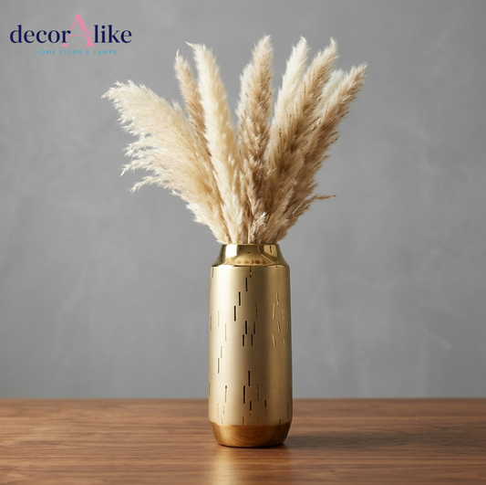 A gold cylindrical vase with a brushed metallic finish and small vertical cutout details, holding dried pampas grass, set on a wooden surface against a textured gray wall. The decorAlike brand logo is visible in the top left corner.