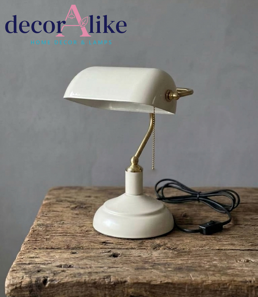 A vintage-style desk lamp with an off-white shade and base, a gold-toned adjustable neck, and a black power cord, displayed on a rustic wooden surface against a gray wall. The 'decorAlike' brand logo is visible in the top left corner.